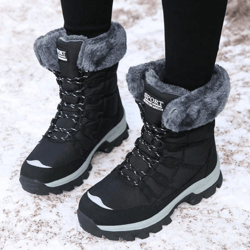 A pair of black faux fur-lined snow boots with grey soles and black and white laces are shown on a model standing in snow.