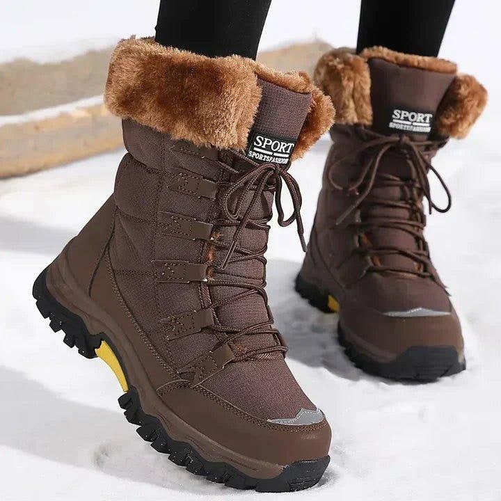 A pair of brown faux fur-lined snow boots with black and yellow soles and brown laces are shown on a model standing in snow.