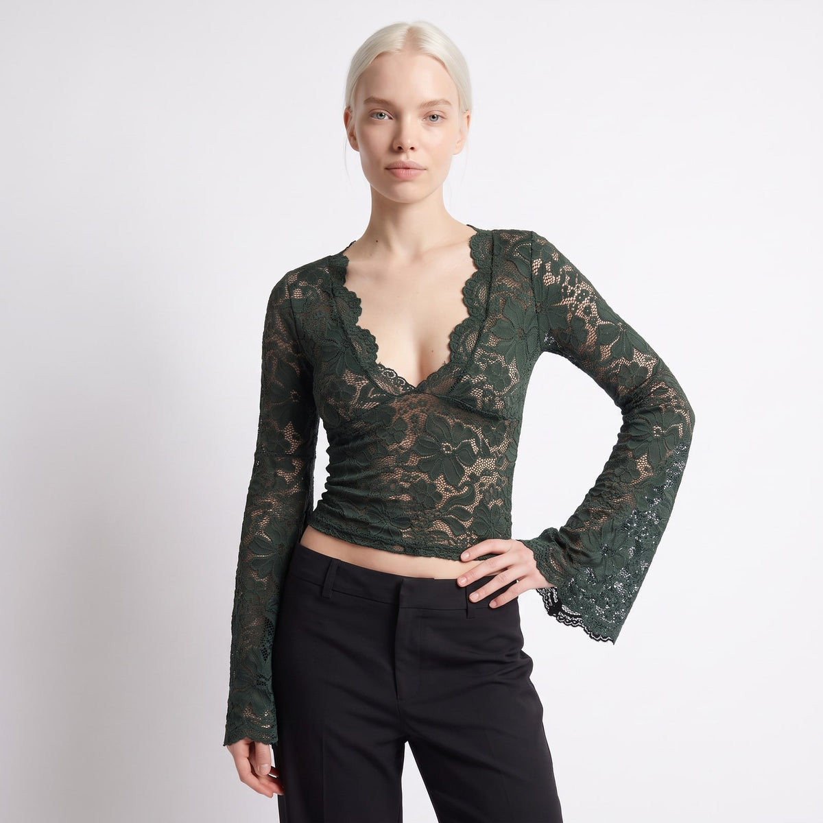 Front view of a woman wearing a dark green floral lace crop top with bell sleeves and black trousers.