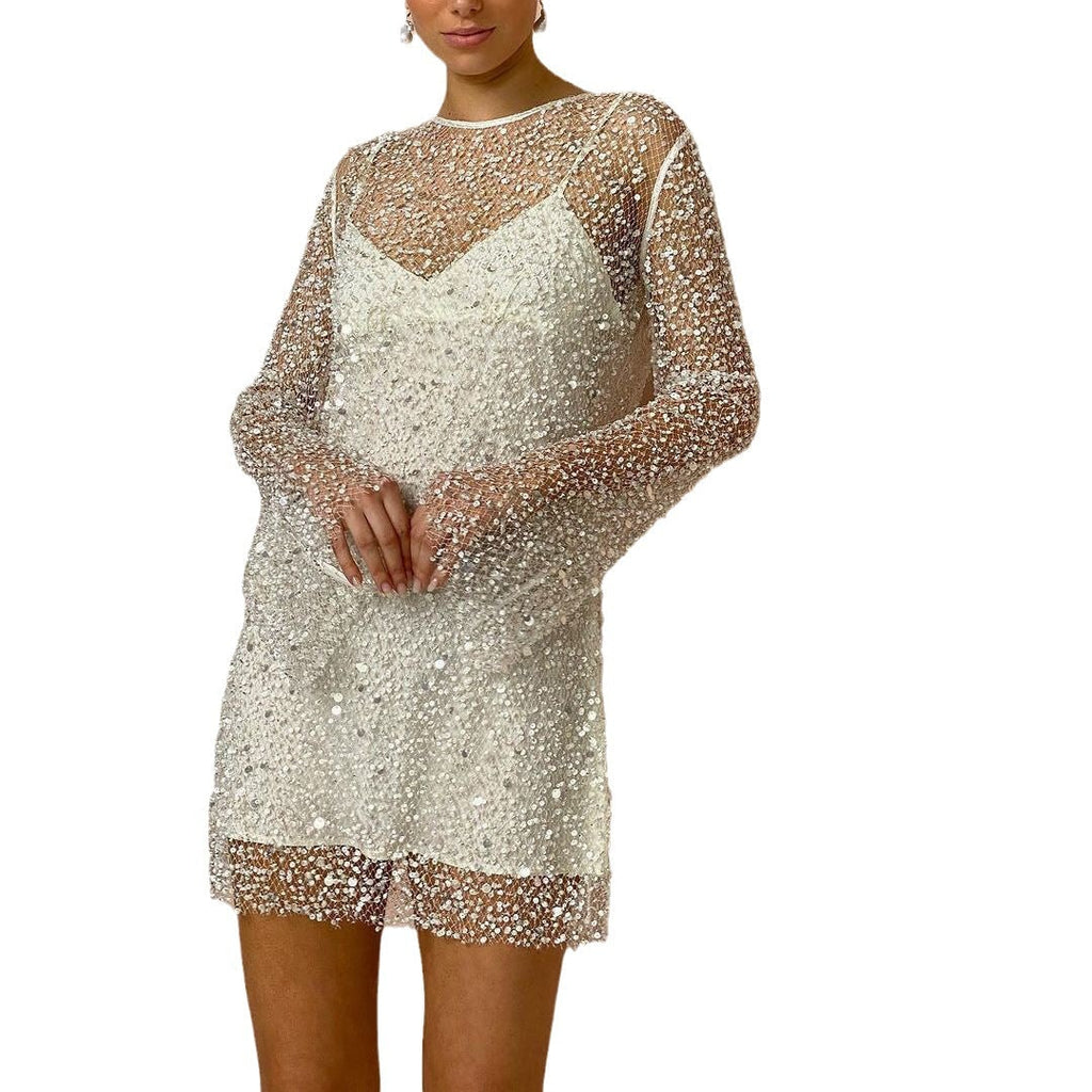 A close-up front view of a model wearing a white sequined sheer long-sleeve mini dress with an inner slip, posing with hands clasped against a white background.
