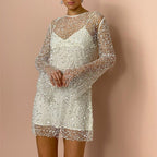 A close-up front view of a model wearing a white sequined sheer long-sleeve mini dress with an inner slip, posing with hands clasped against a pink background.