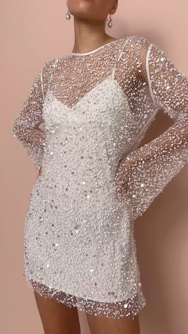 A close-up front view of a model wearing a white sequined sheer long-sleeve mini dress with an inner slip, posing with hands on hips against a pink background.