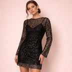 A full-body front view of a model wearing a black sequined sheer long-sleeve mini dress with an inner slip, looking at the camera against a pink background.