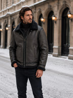 Front view of a man wearing a dark grey shearling aviator jacket with the zipper closed, standing on a snowy street.