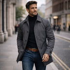 A male model wearing a dark grey mac coat over a black turtleneck and blue jeans, looking to the side on a city street.