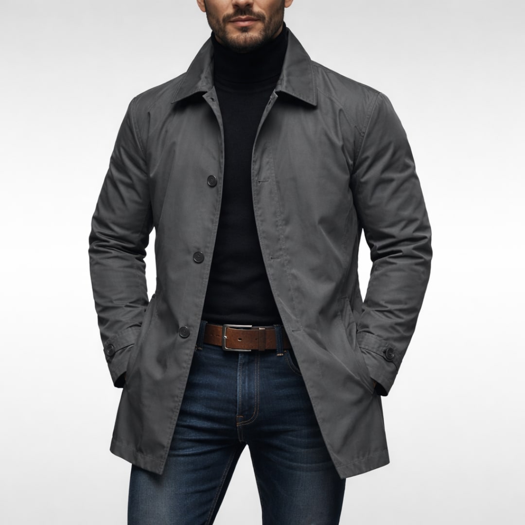 A male model wearing a dark grey mac coat over a black turtleneck and blue jeans, posing front-facing against a white background.