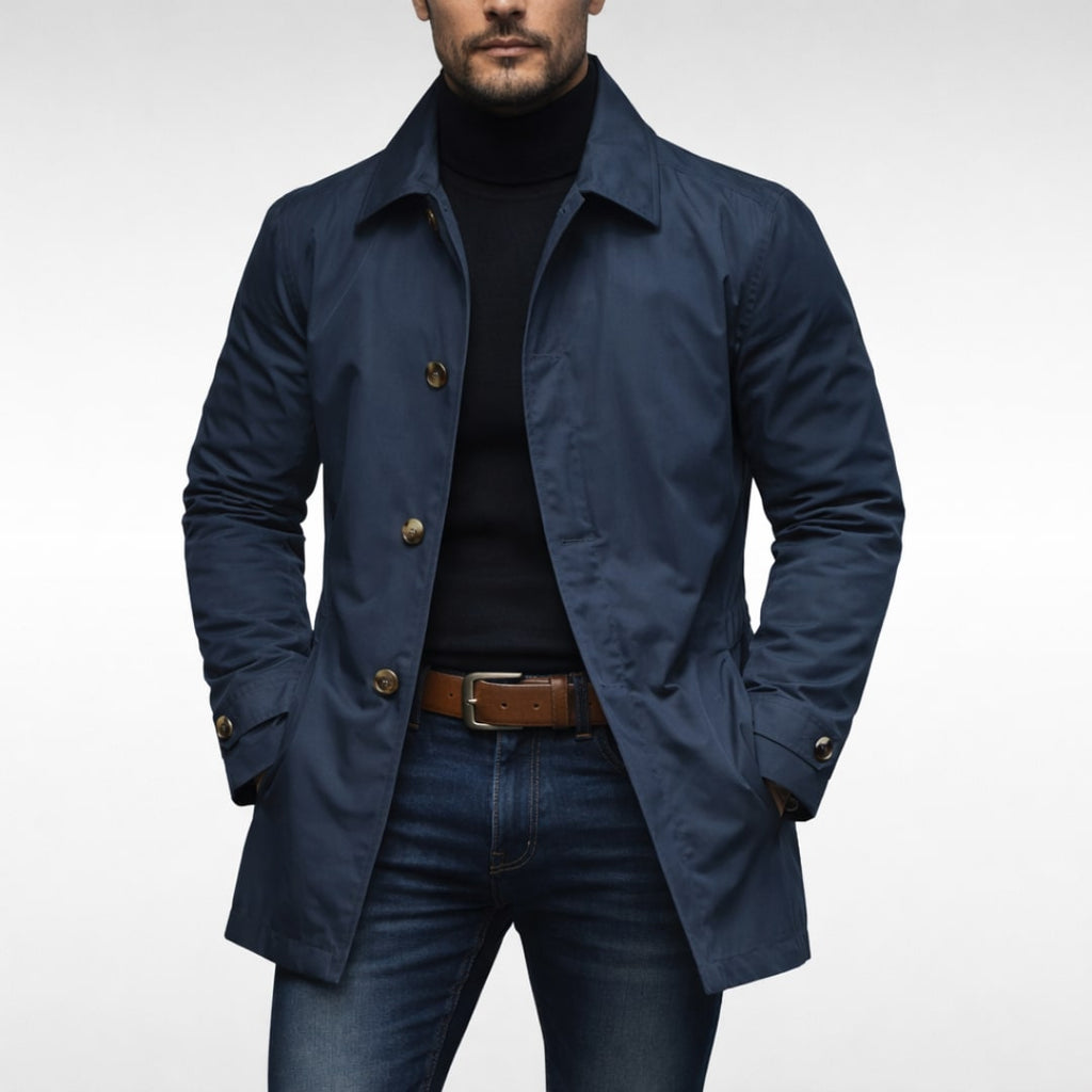 A male model wearing a navy blue mac coat over a black turtleneck and blue jeans, posing front-facing against a white background.