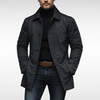 A male model wearing a dark navy mac coat over a black turtleneck and blue jeans, posing front-facing against a white background.