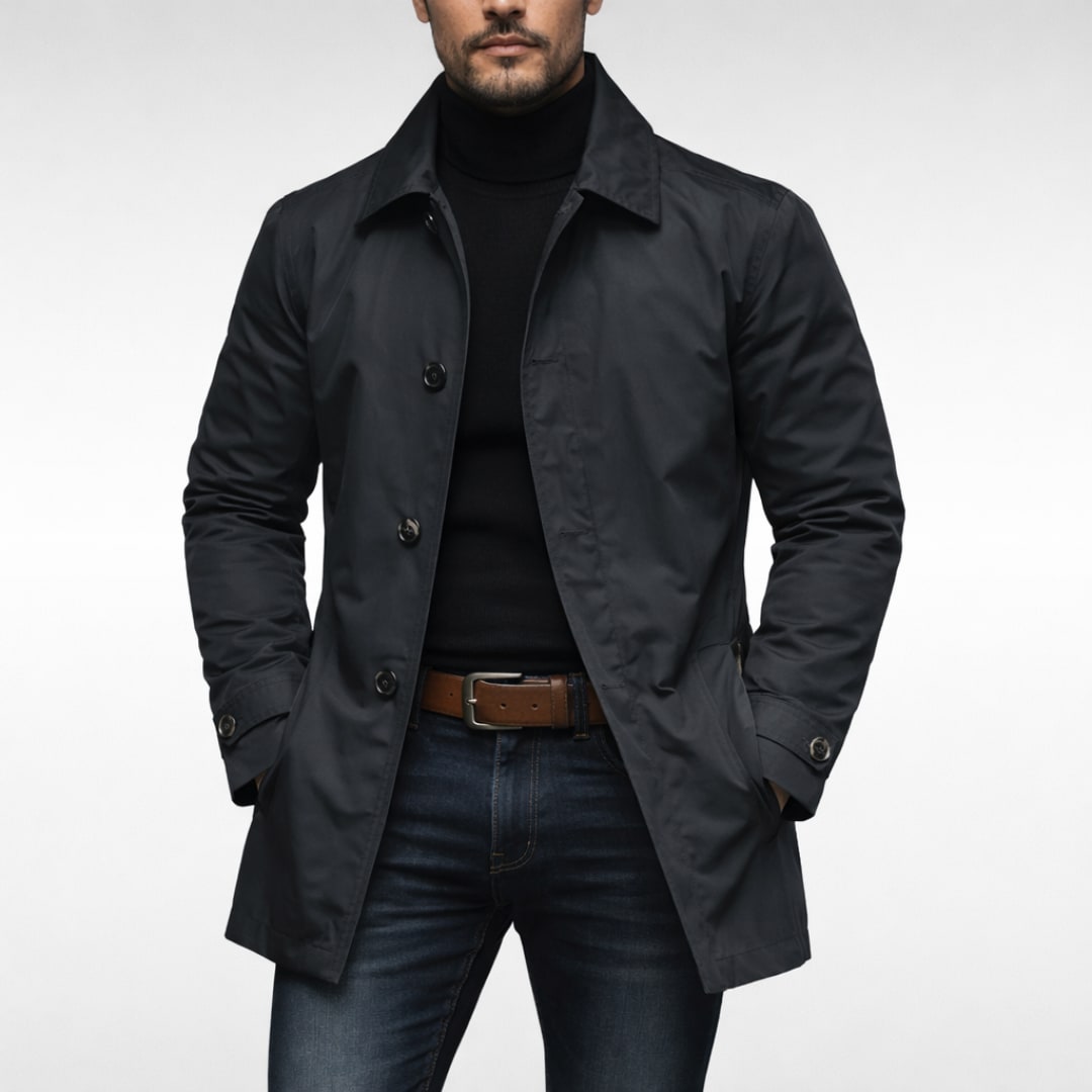 A male model wearing a dark navy mac coat over a black turtleneck and blue jeans, posing front-facing against a white background.