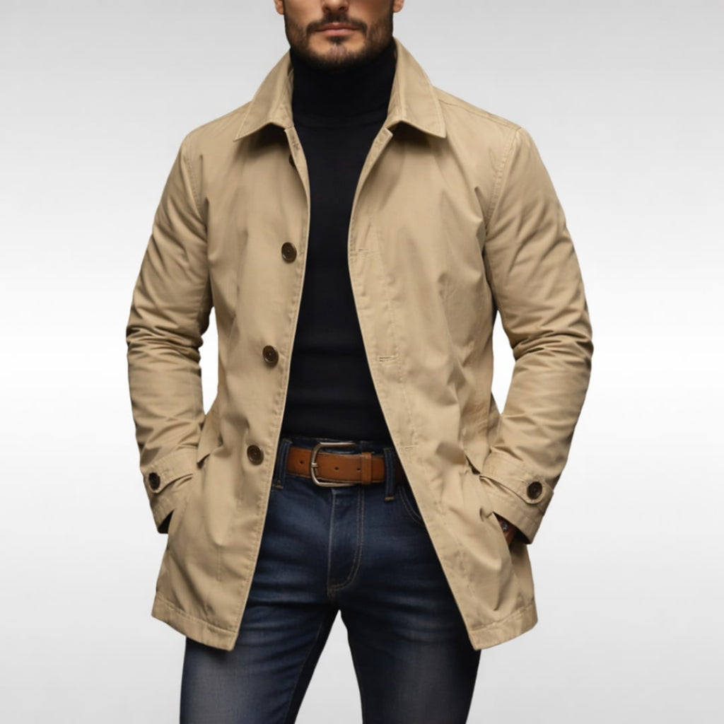 A male model wearing a beige mac coat over a black turtleneck and blue jeans, posing front-facing against a white background.