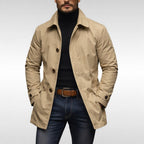 A male model wearing a beige mac coat over a black turtleneck and blue jeans, posing front-facing against a white background.