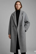 Front view of a woman wearing the grey double-breasted long coat open over a black turtleneck and trousers.