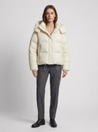 Front view of a woman wearing a cream-colored puffer jacket, grey trousers, and black boots, standing against a white background.