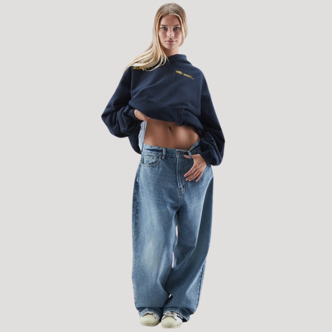 Front view of a female model wearing a dark blue cropped oversized hoodie with a yellow chest logo and baggy light wash jeans.
