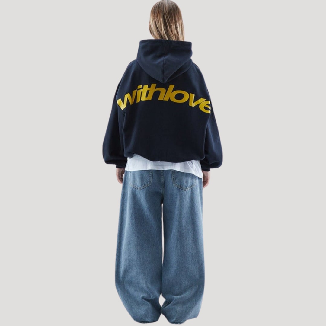 Back view of a female model wearing a dark blue cropped oversized hoodie with a large yellow 'withlove' graphic and baggy light wash jeans.