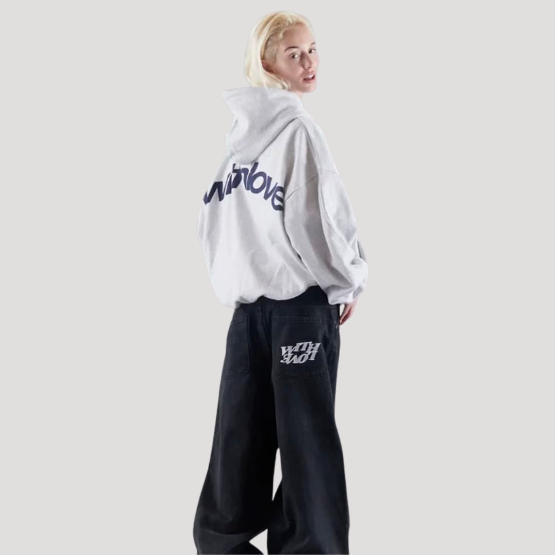 Side-back view of a female model wearing a light grey oversized hoodie with a large dark 'withlove' graphic and baggy black jeans.