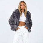 Front view of a woman wearing a dark grey faux fur jacket over a white crop top and white pants.