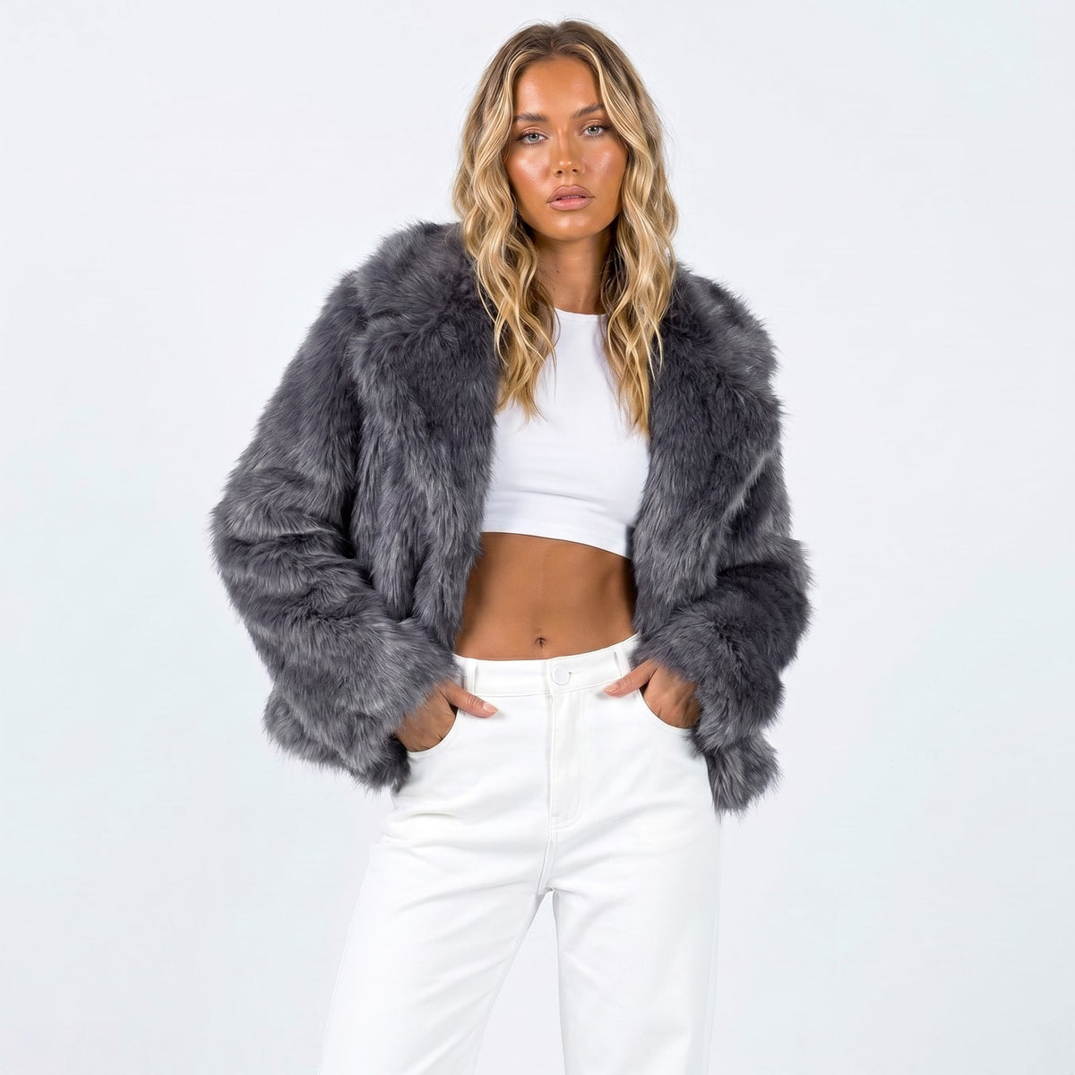 Front view of a woman wearing a dark grey faux fur jacket over a white crop top and white pants.