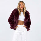 A woman models a burgundy faux fur jacket, white crop top, and white pants, posing with hands in pockets.
