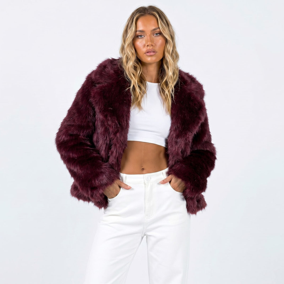 A woman models a burgundy faux fur jacket, white crop top, and white pants, posing with hands in pockets.
