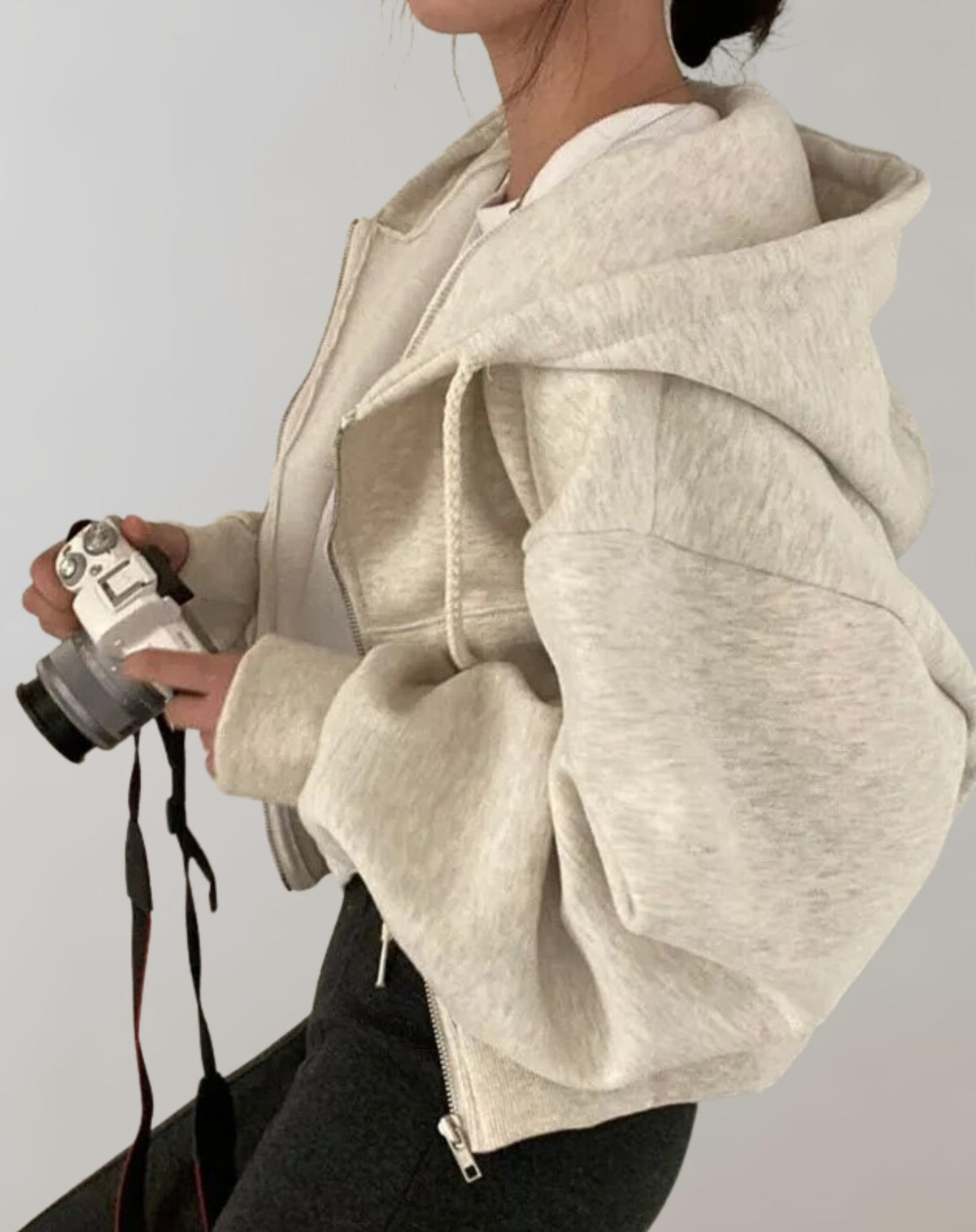 Side view of a model wearing a light grey marl cropped oversized zip-up hoodie, holding a camera.
