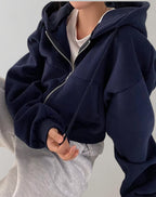 Front view of a model sitting, wearing a navy cropped oversized zip-up hoodie over a white t-shirt and light grey sweatpants.