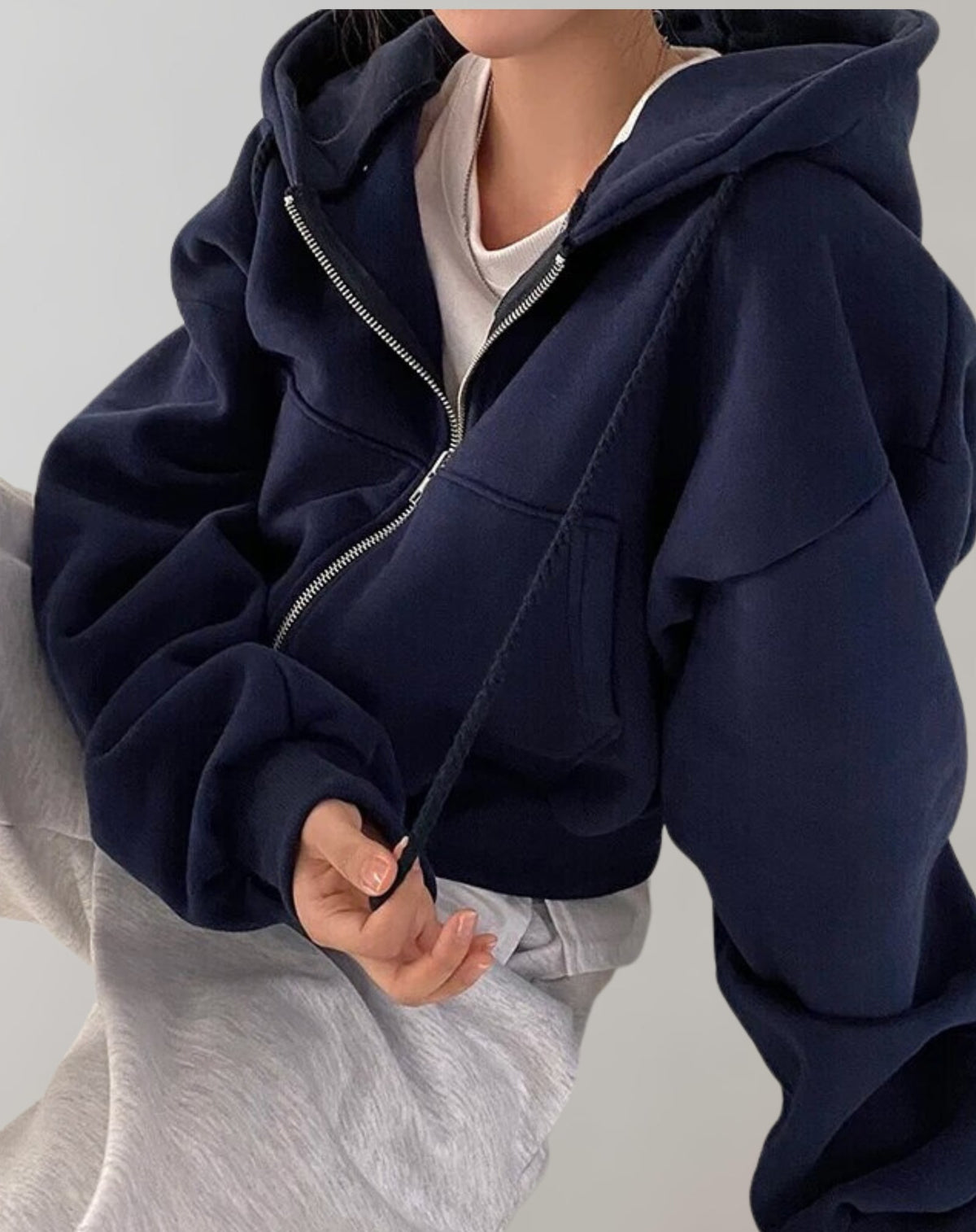 Front view of a model sitting, wearing a navy cropped oversized zip-up hoodie over a white t-shirt and light grey sweatpants.