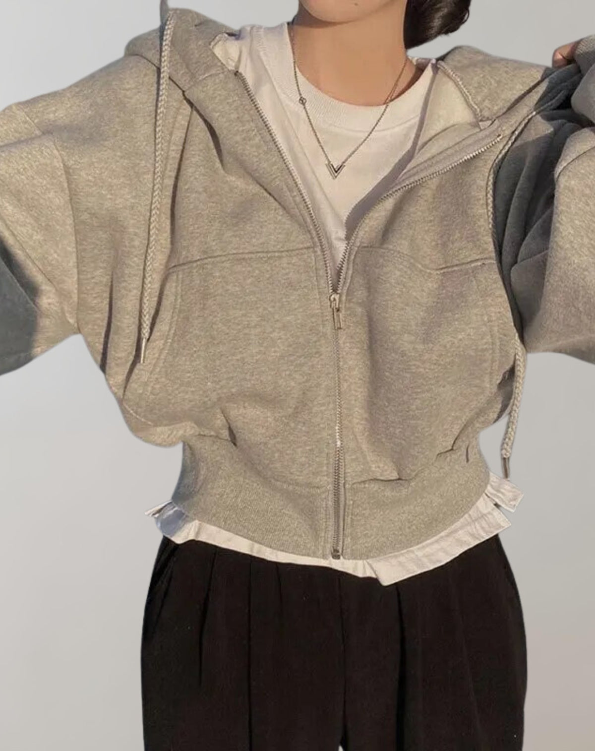 Front view of a model wearing a grey cropped oversized zip-up hoodie over a white t-shirt and black pleated pants.