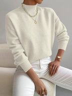 Front view of a woman wearing a cream mock neck sweater with white pants, accessorized with a gold necklace and watch.