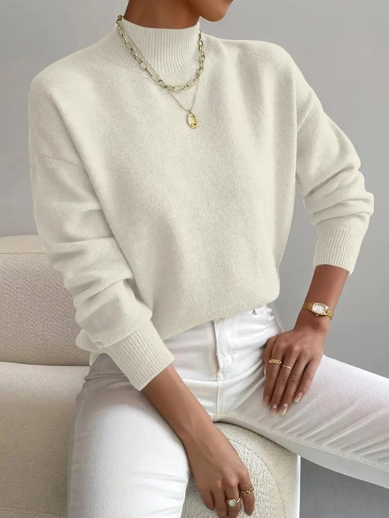 Front view of a woman wearing a cream mock neck sweater with white pants, accessorized with a gold necklace and watch.