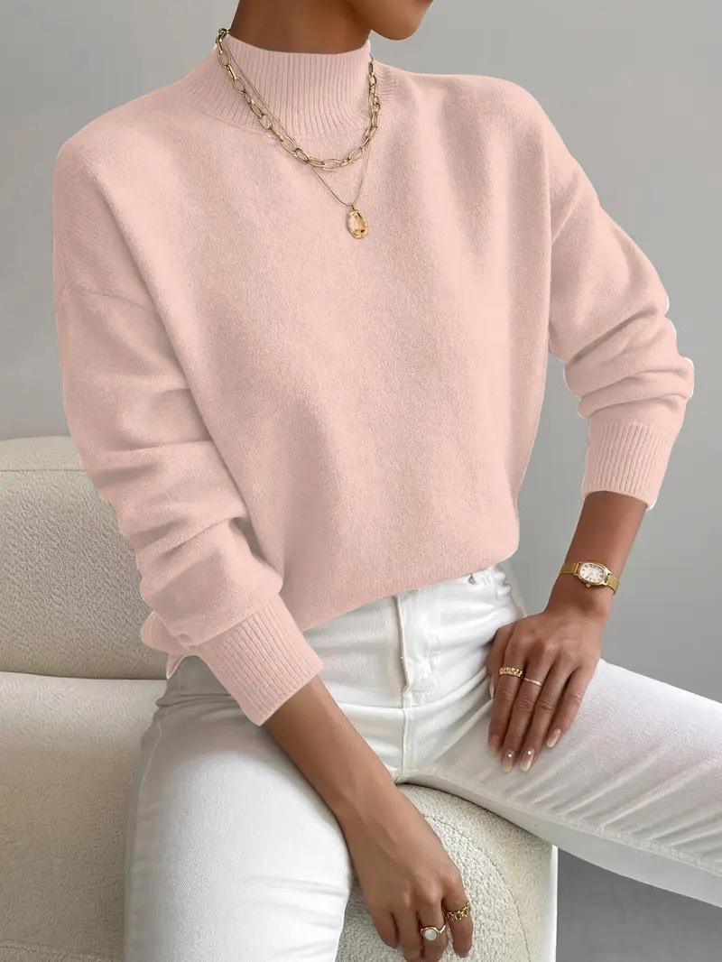 Front view of a woman wearing a light pink mock neck sweater with white pants, accessorized with a gold necklace and watch.