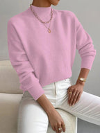 Front view of a woman wearing a bright pink mock neck sweater with white pants, accessorized with a gold necklace and watch.
