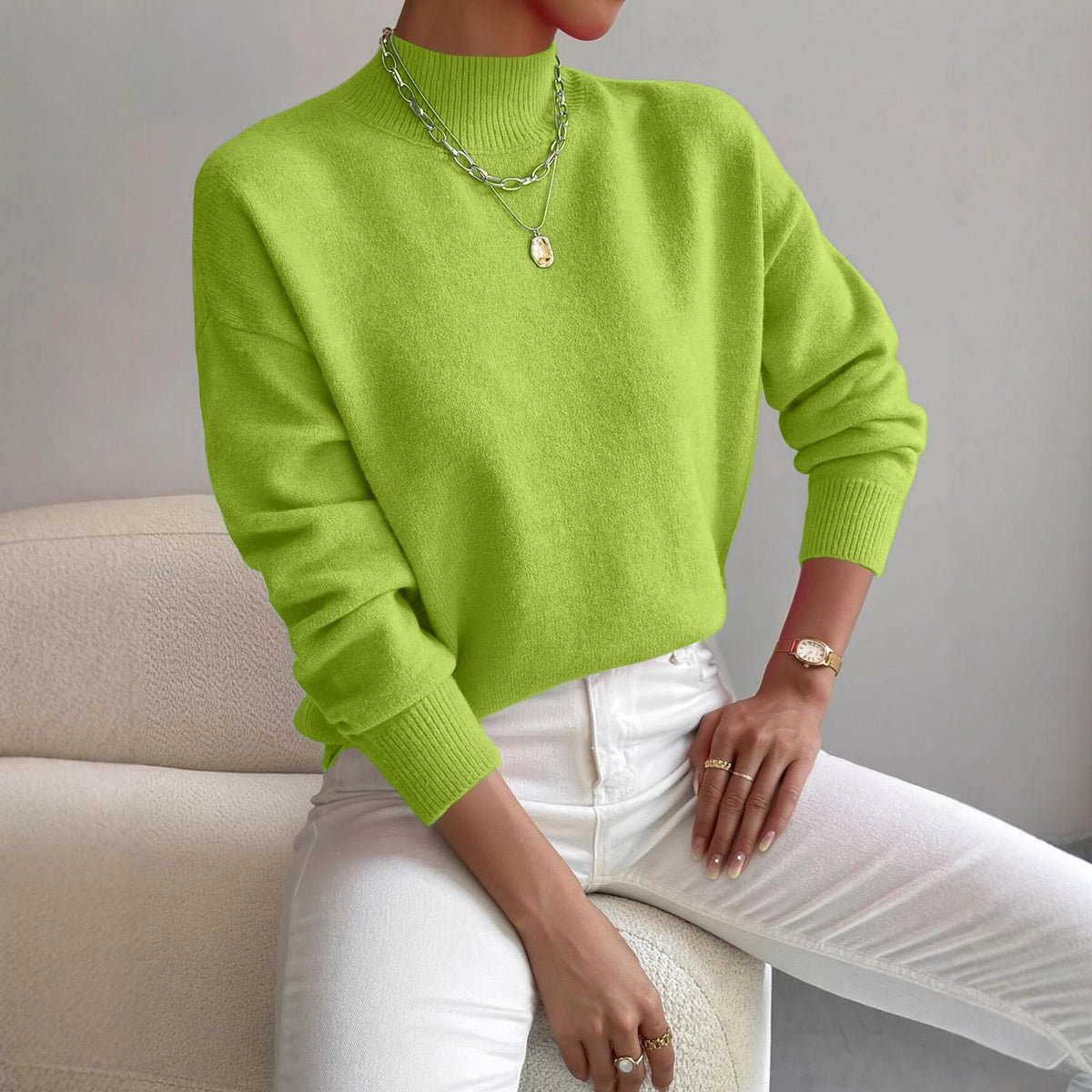 Front view of a woman wearing a neon green mock neck sweater with white pants, accessorized with a silver necklace and gold watch.