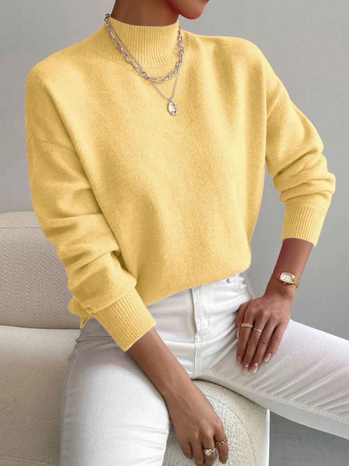Front view of a woman wearing a yellow mock neck sweater with white pants, accessorized with a silver necklace and gold watch.