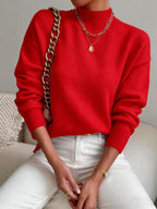 Front view of a woman wearing a red mock neck sweater with white pants, accessorized with a gold necklace, watch, and a gold chain strap bag.