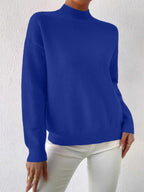 Full front view of a woman wearing a royal blue mock neck sweater with white pants, standing against a white textured wall.