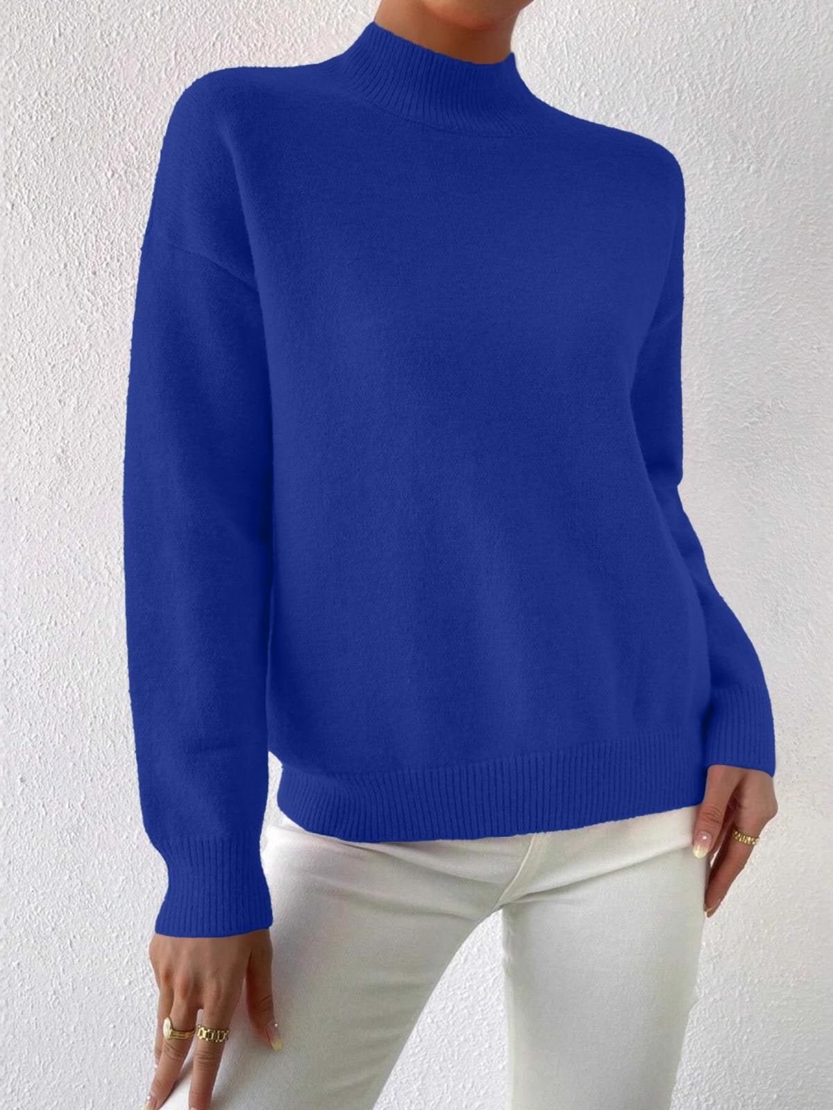 Full front view of a woman wearing a royal blue mock neck sweater with white pants, standing against a white textured wall.