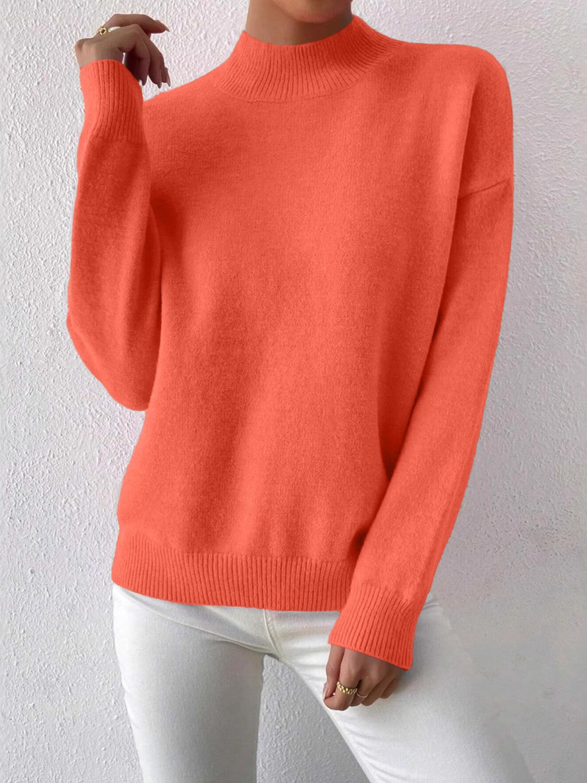 Full front view of a woman wearing an orange mock neck sweater with white pants, standing against a white textured wall.