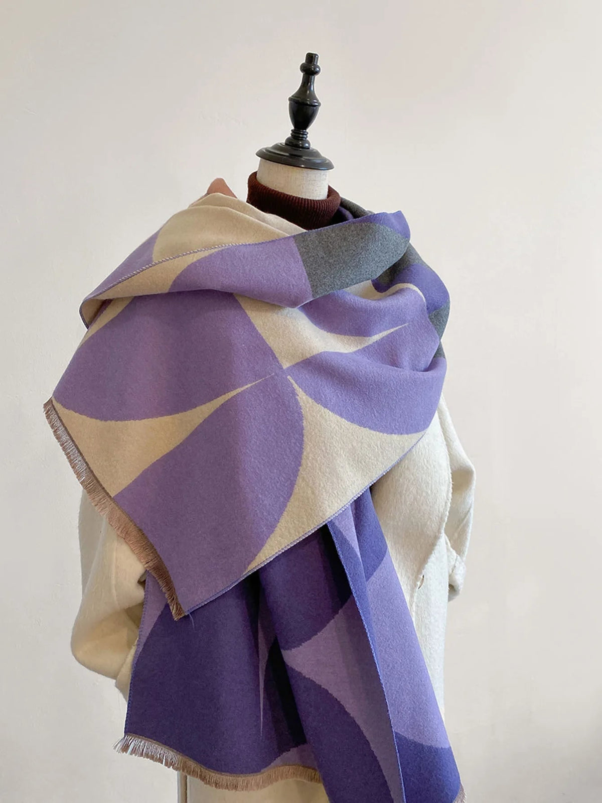 Front view of a purple, cream, and grey geometric pattern blanket scarf draped on a mannequin wearing a cream coat.