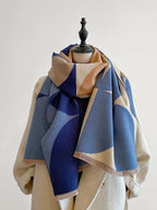 Front view of a blue, brown, and cream geometric pattern blanket scarf draped on a mannequin wearing a cream coat.