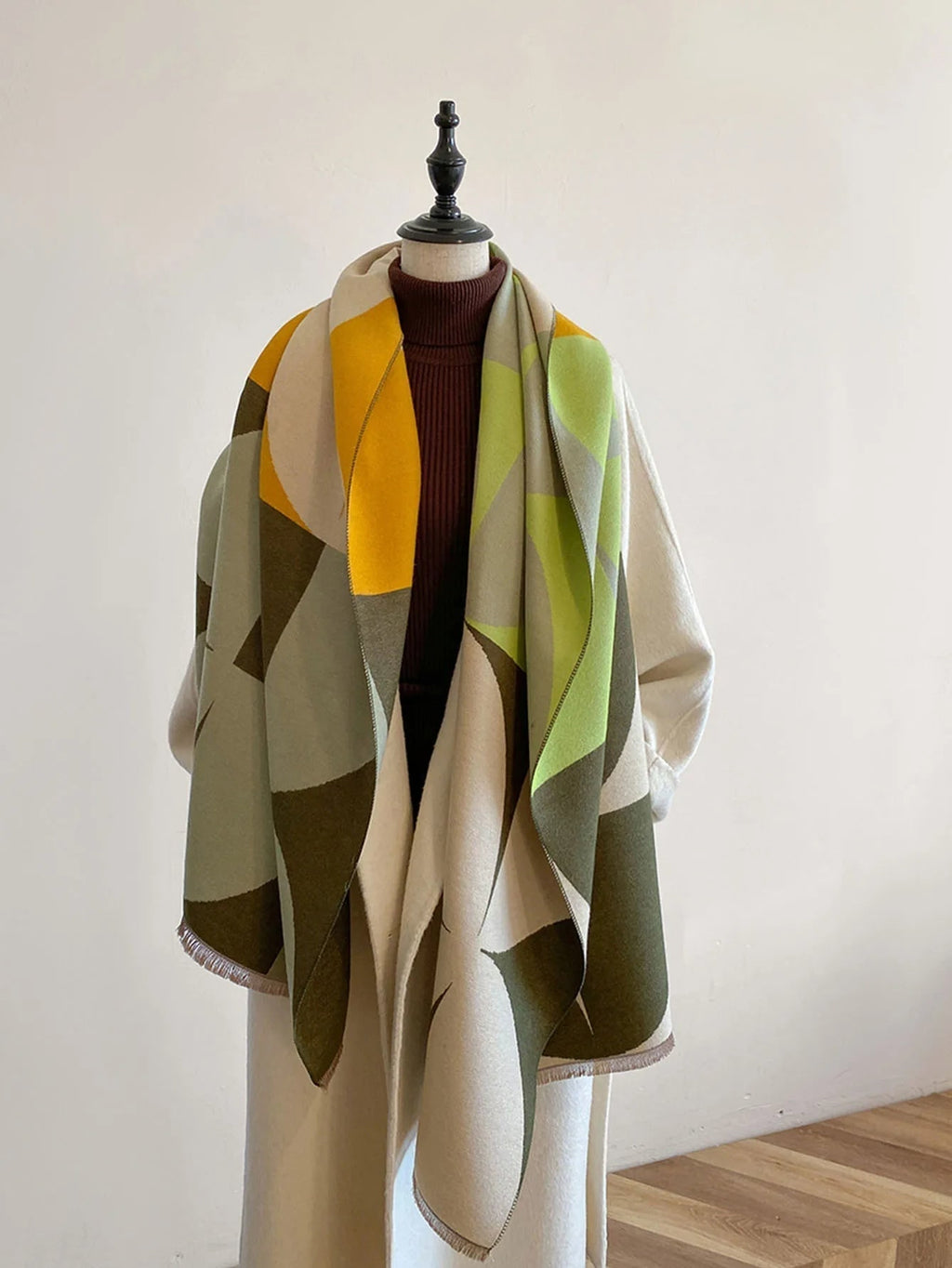 Front view of a yellow, green, and grey geometric pattern blanket scarf draped on a mannequin wearing a cream coat.