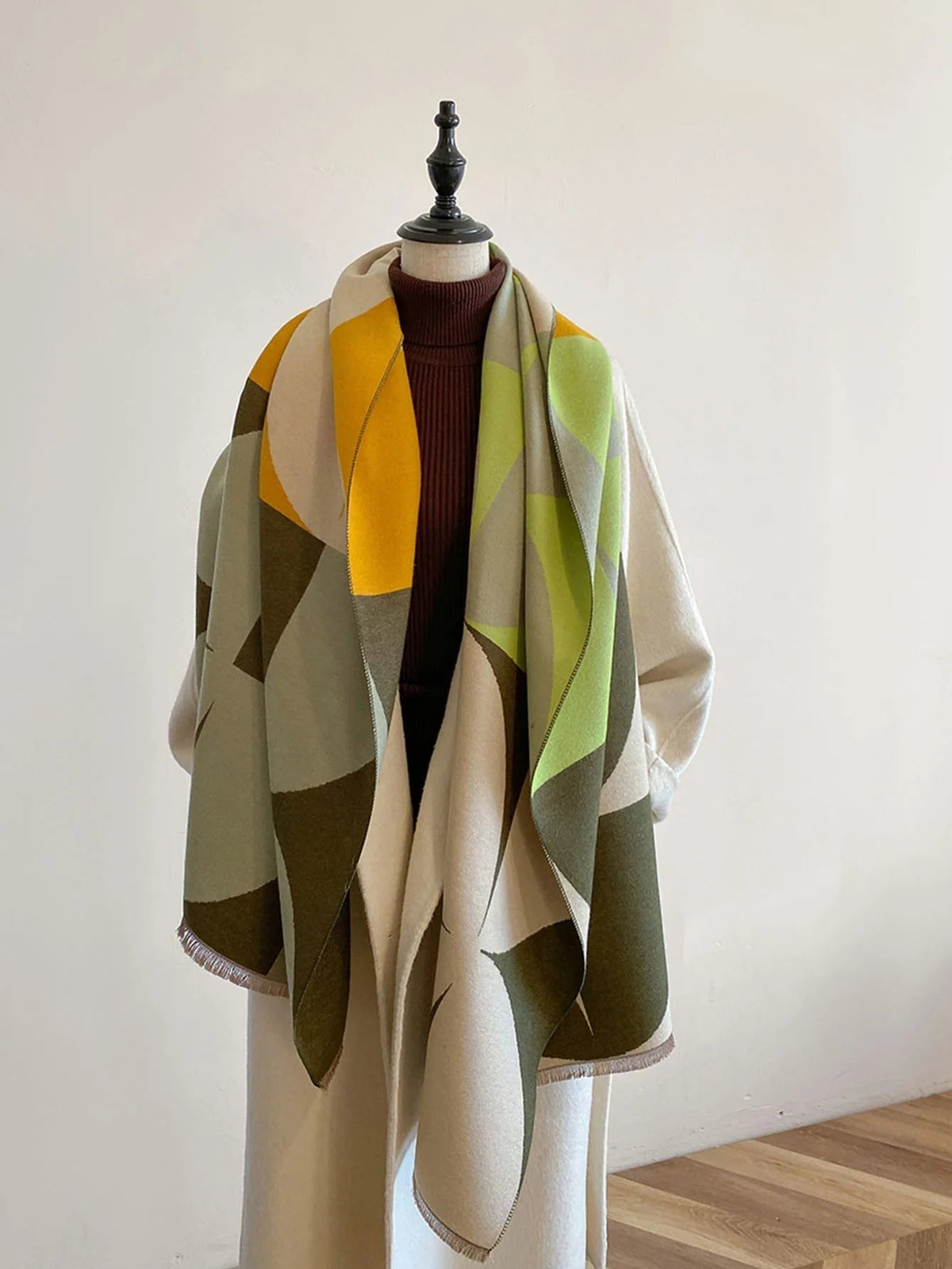 Front view of a yellow, green, and grey geometric pattern blanket scarf draped on a mannequin wearing a cream coat.
