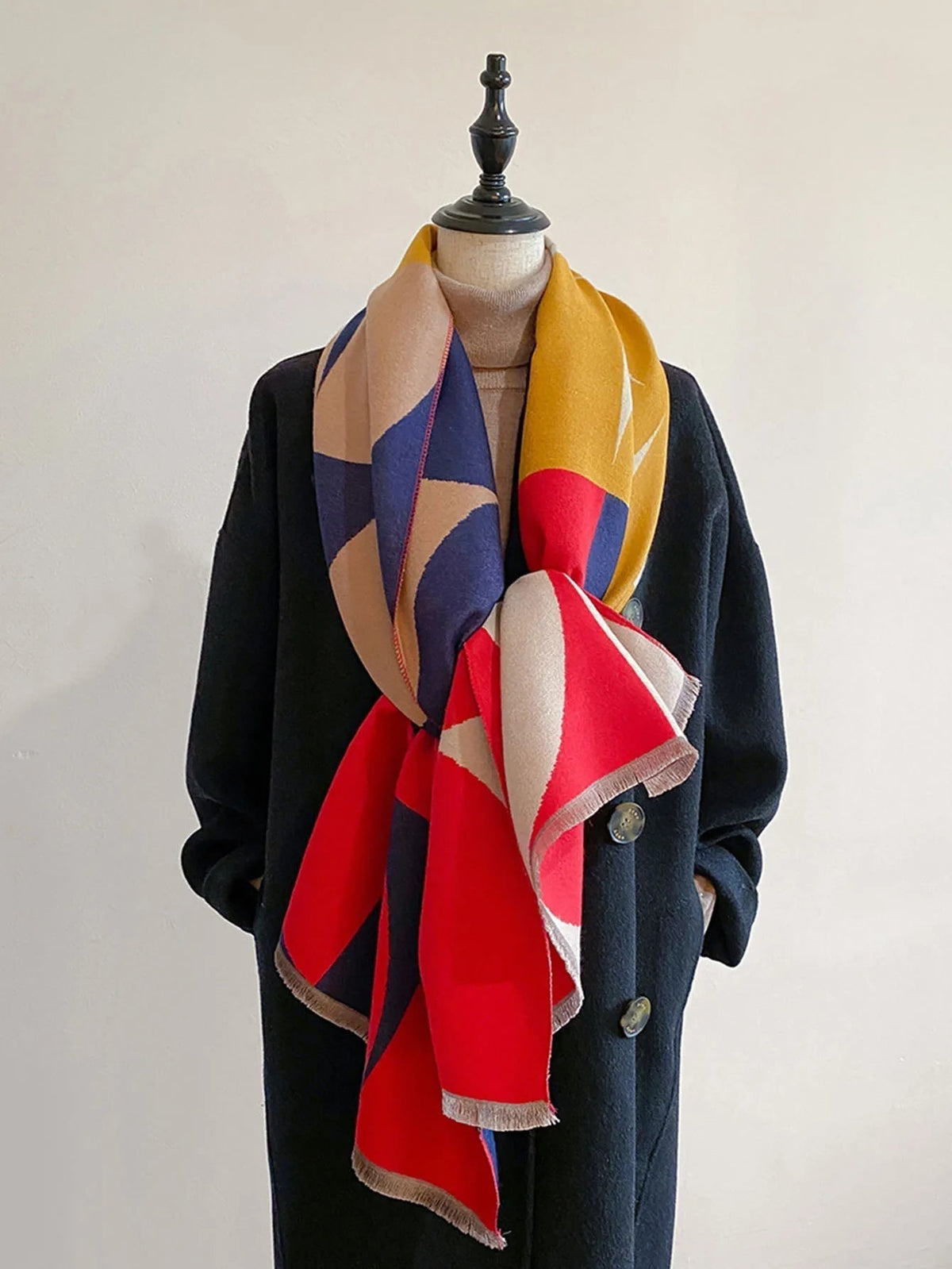 Front view of a red, yellow, blue, and cream geometric pattern blanket scarf tied on a mannequin wearing a dark coat.