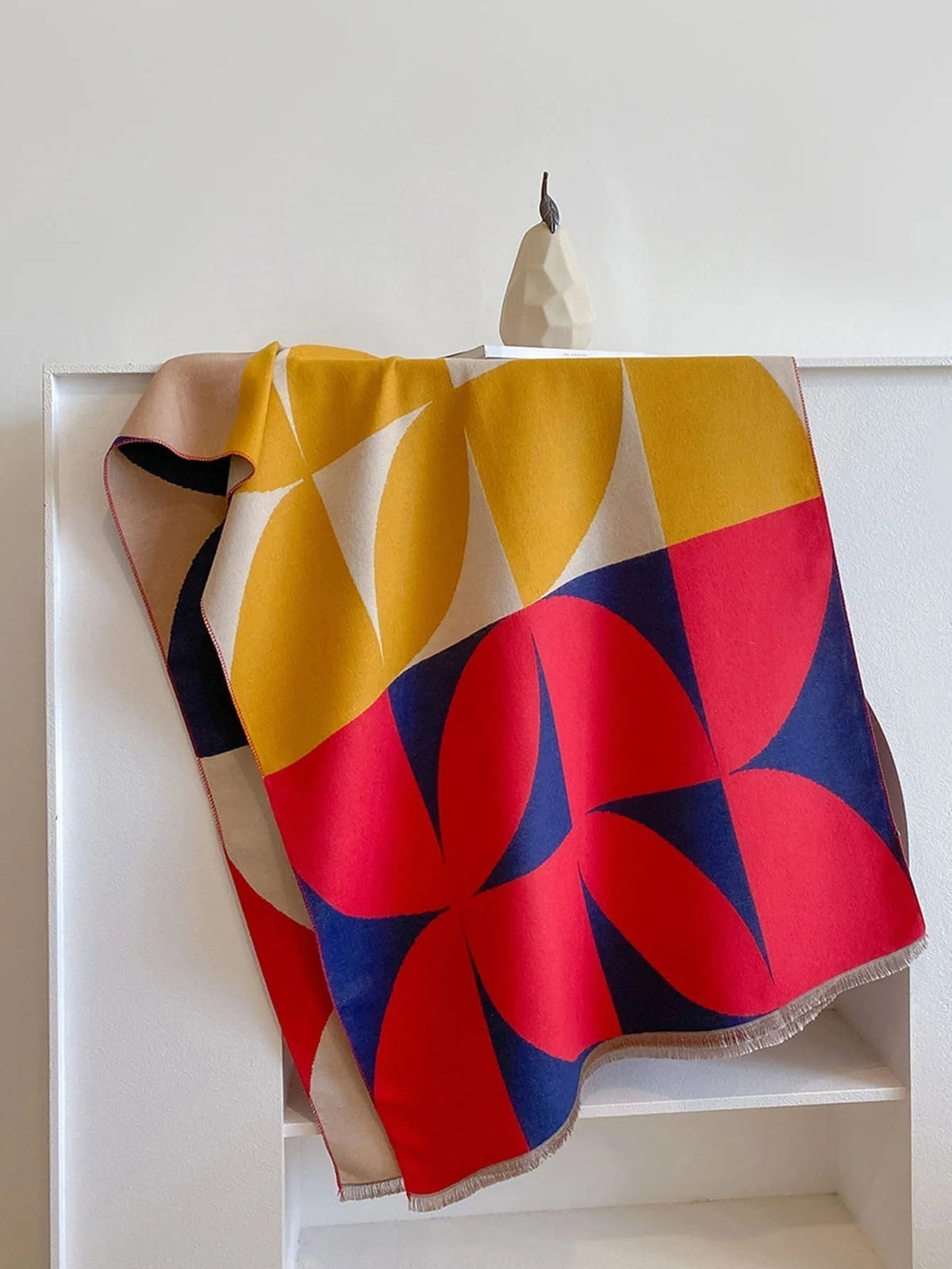 Overhead view of a red, yellow, blue, and cream geometric pattern blanket scarf folded on a white shelf.