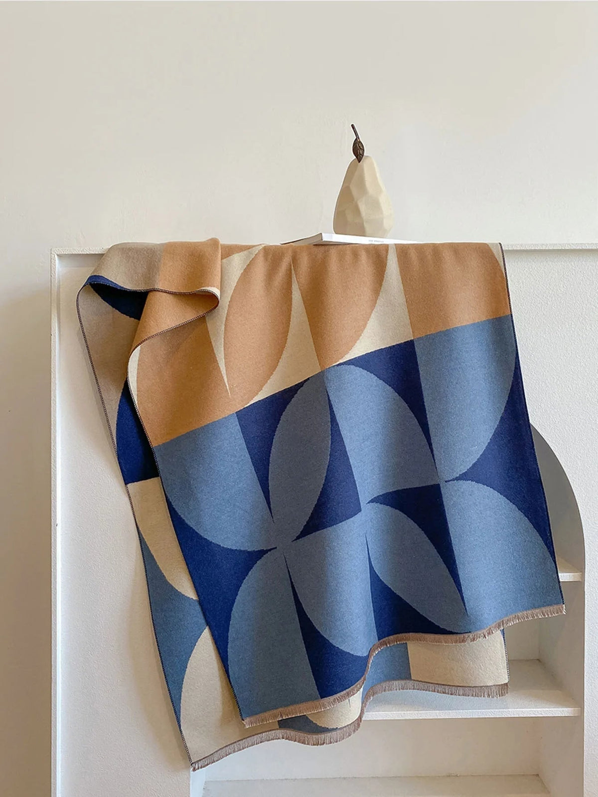 Overhead view of a blue, brown, and cream geometric pattern blanket scarf folded on a white shelf.