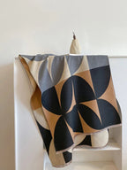 Overhead view of a black, brown, cream, and grey geometric pattern blanket scarf folded on a white shelf.