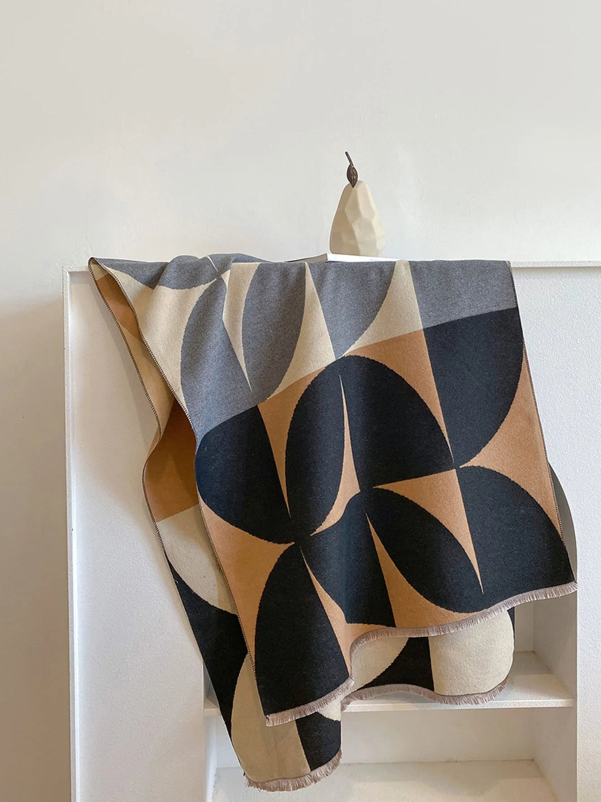 Overhead view of a black, brown, cream, and grey geometric pattern blanket scarf folded on a white shelf.