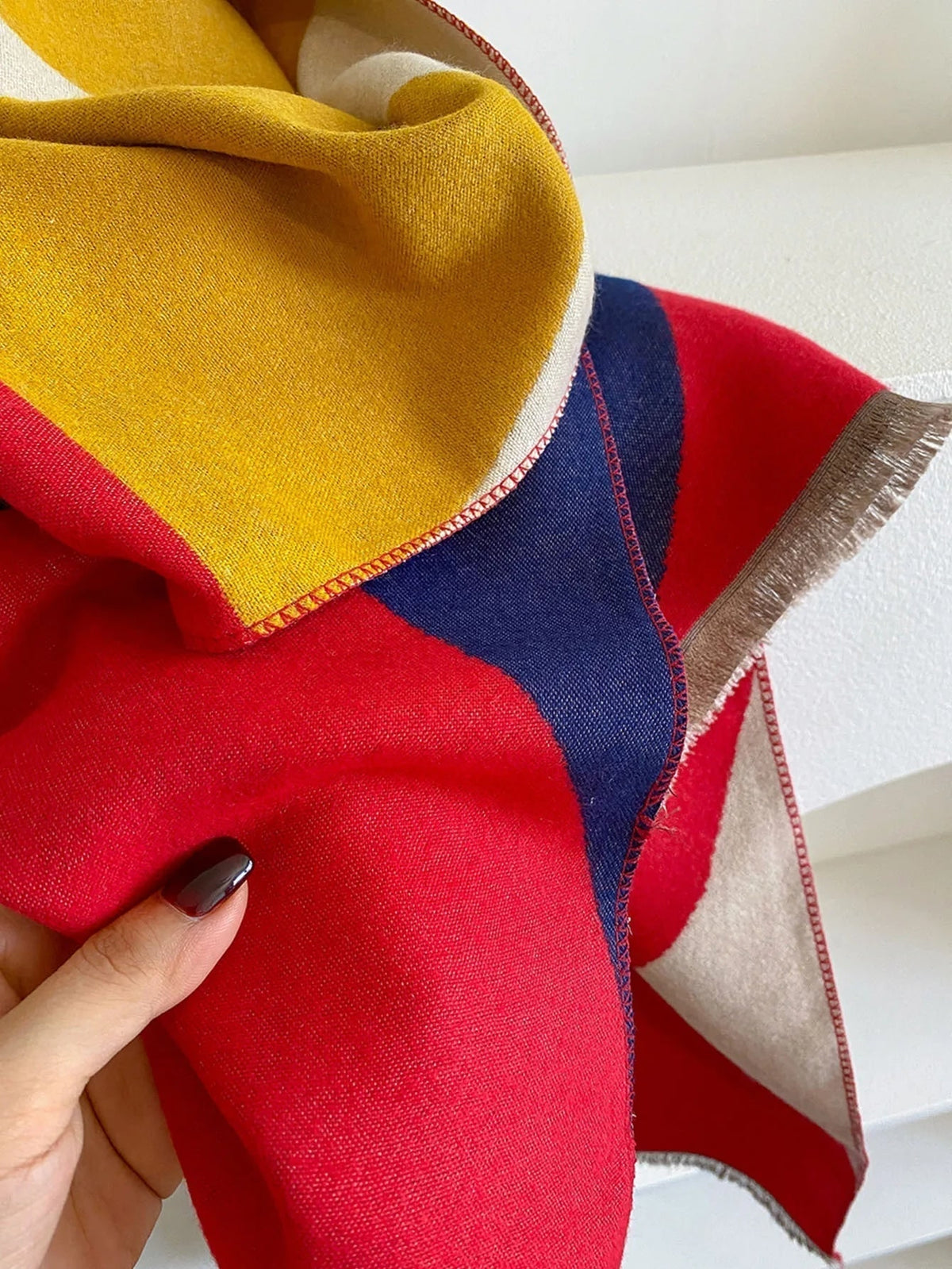 A close-up shot of a red, yellow, navy blue, and cream geometric patterned scarf with fringed edges, held by a hand.