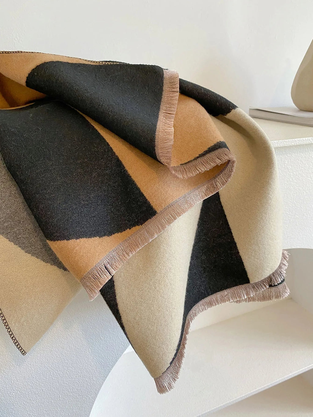 A black, tan, and cream geometric patterned scarf with fringed edges, draped over a white surface.