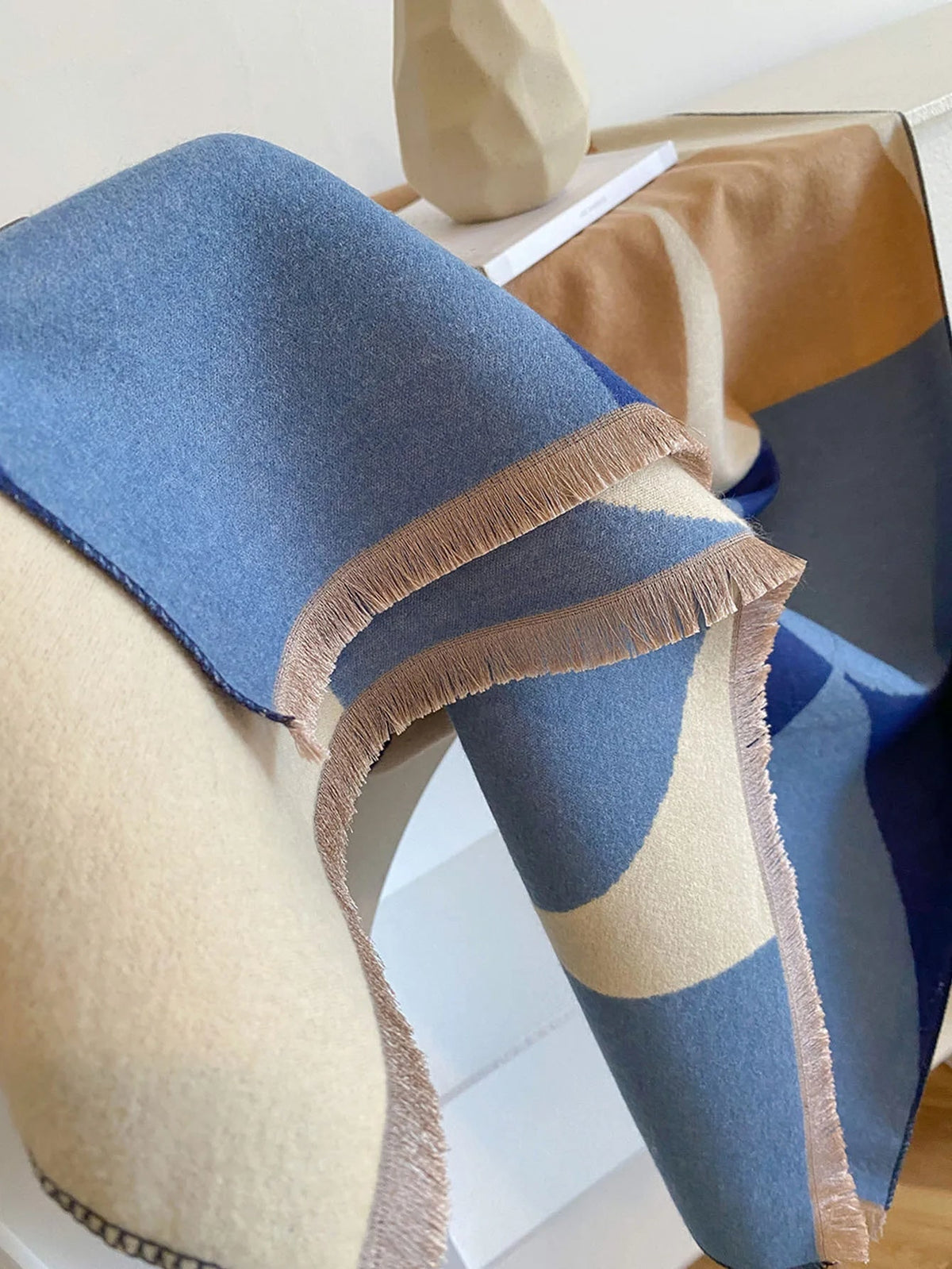A blue, light blue, tan, and cream geometric patterned scarf with fringed edges, draped over a white surface.